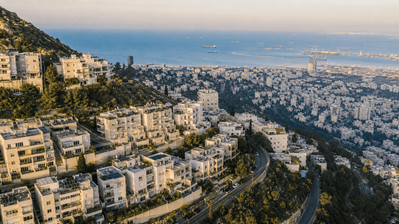 תצלום אווירי של חיפה. בניינים מודרניים יורדים במורד הכרמל לעבר המפרץ, ים תיכון ברקע, שעת זהב