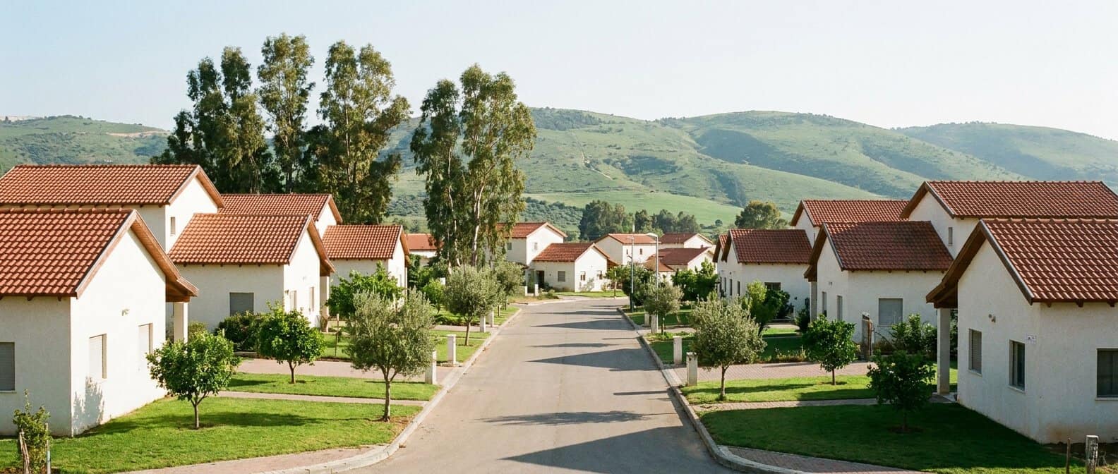 שכונת הרחבה בקיבוץ עם בתים פרטיים חדשים בגגות רעפים, מדשאות ועצים, ורכסי הרים ירוקים ברקע באור יום